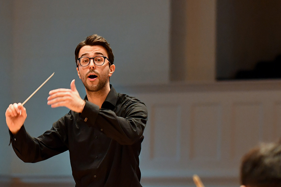 A male student waving his conductors baton, conducting an orchestra rehearsal.